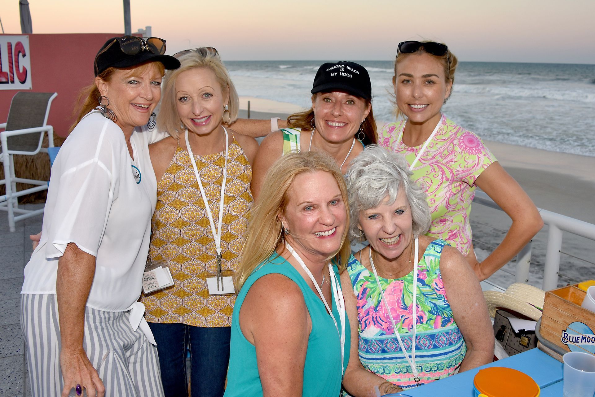 Six women smiling, posing for photo on beach. Wearing summer attire, near building.
