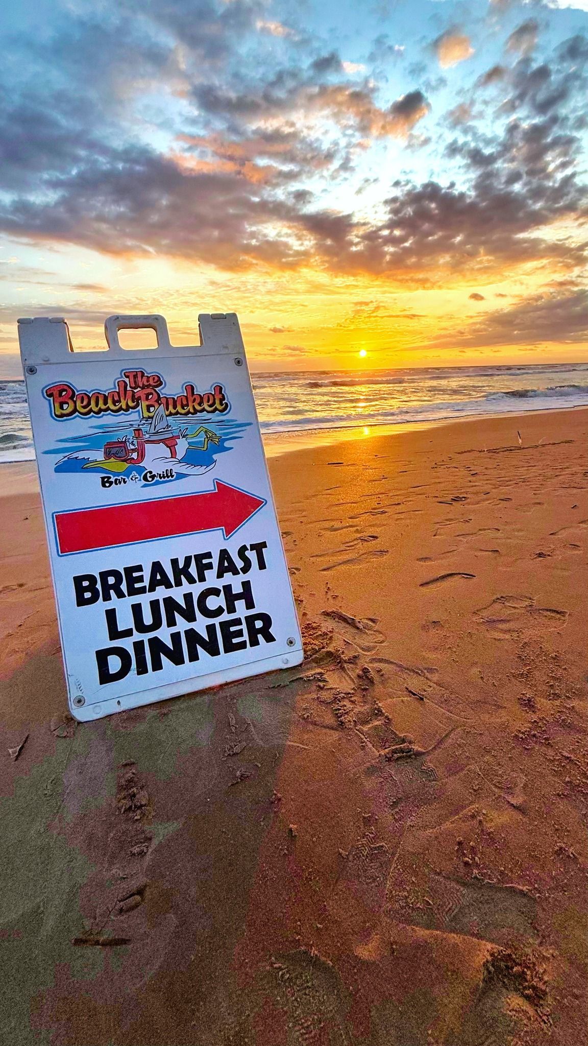 Sign on a beach at sunset with The Beach Bucket Bar and Grill logo with a red arrow advertising breakfast, lunch, and dinner.