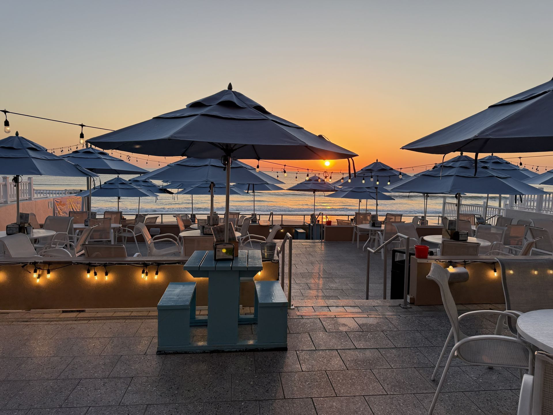 Beachfront restaurant at sunset with blue umbrellas, tables, and ocean view.