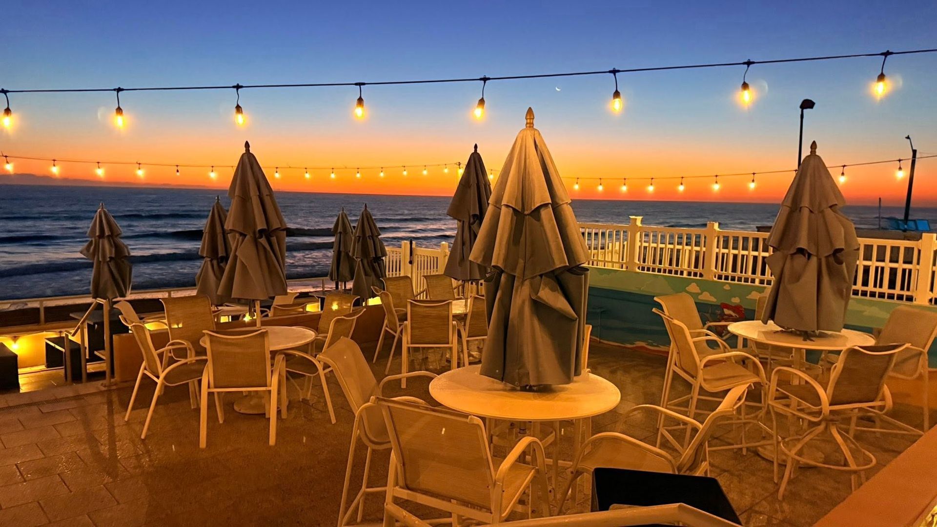 Restaurant patio overlooking the ocean at sunset; tables, umbrellas, string lights.