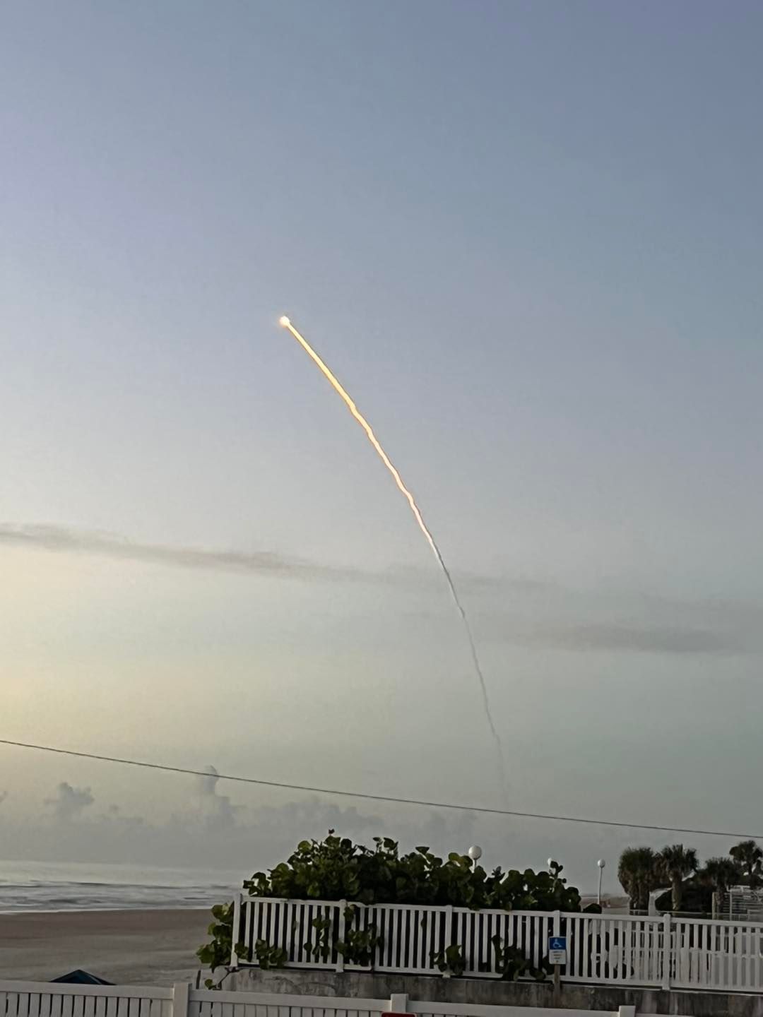 Rocket launching into a light blue sky over a beach, leaving a bright contrail.