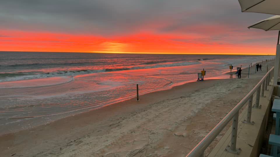 Beach sunset with orange and red sky, ocean waves, and people walking along the shore.