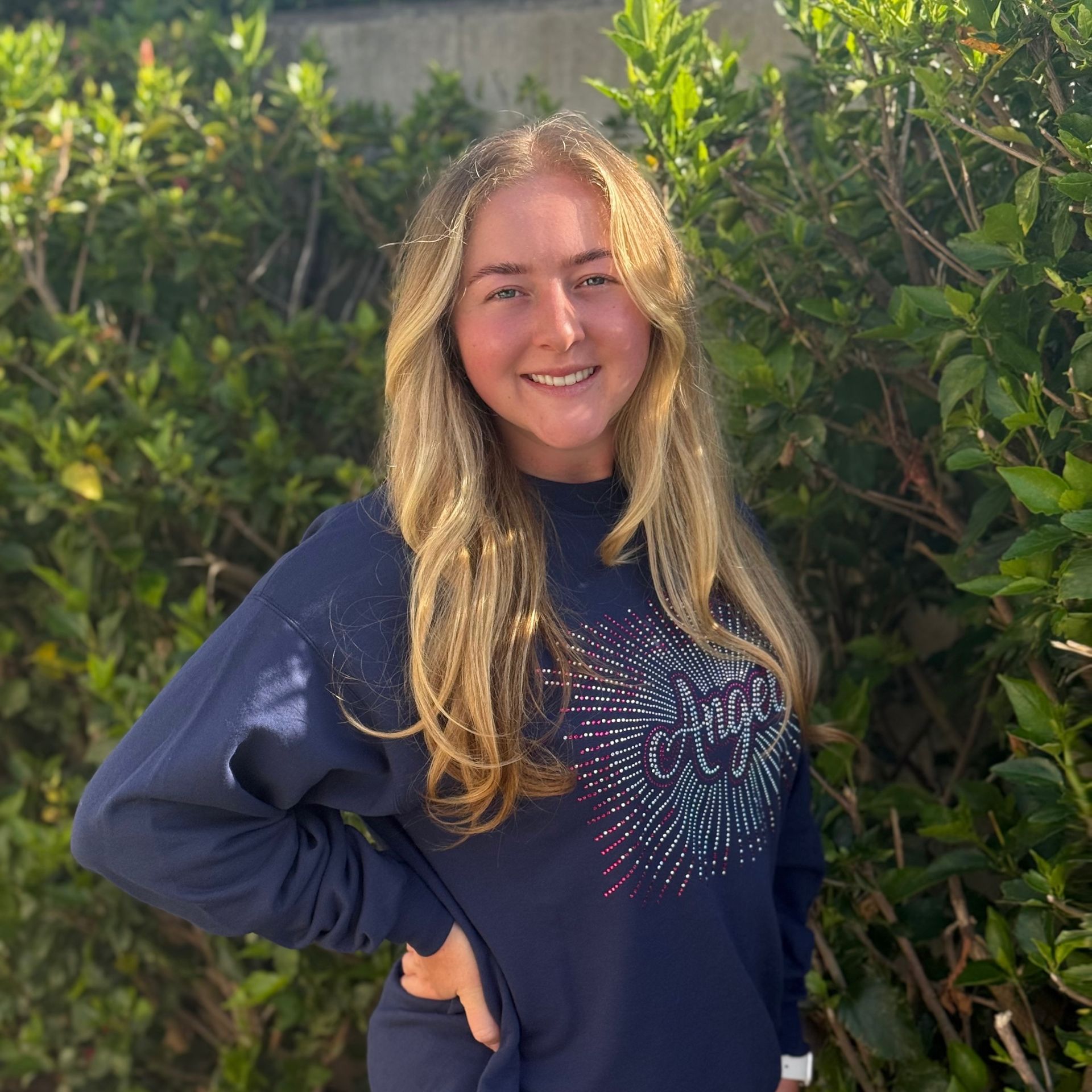 Blond woman smiles outdoors, wearing a blue sweatshirt, hand on hip. Image of Coach Makayla.