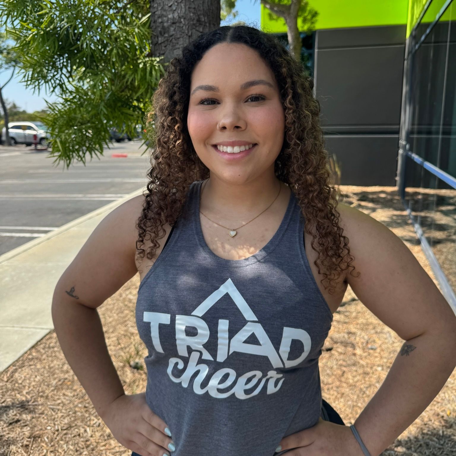 Woman with curly hair wearing a Triad Cheer tank top, hands on hips, smiling outside. Image of Coach Piper.