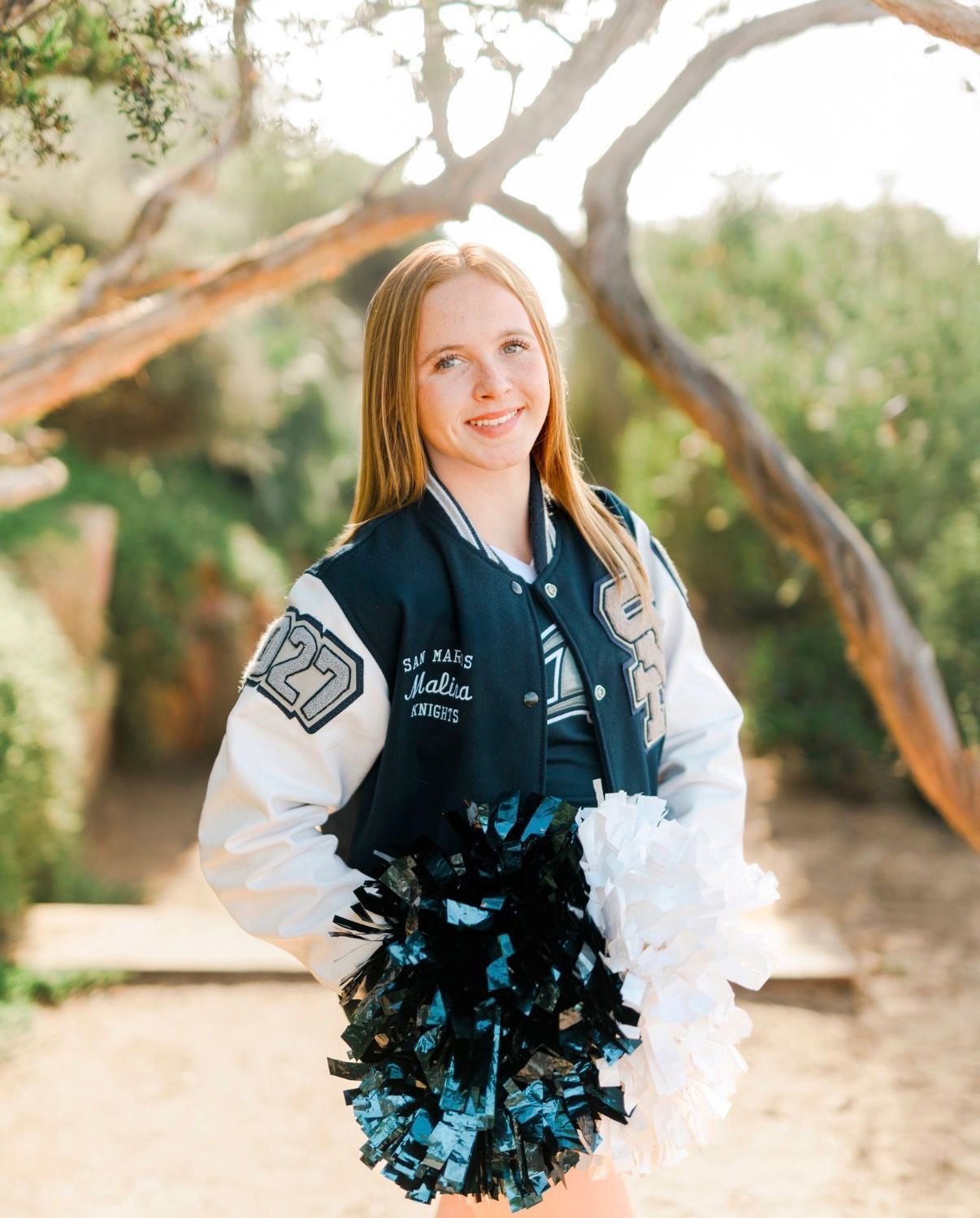 Cheerleader in letterman jacket, holding pom-poms, smiling. Outdoors, trees in background.