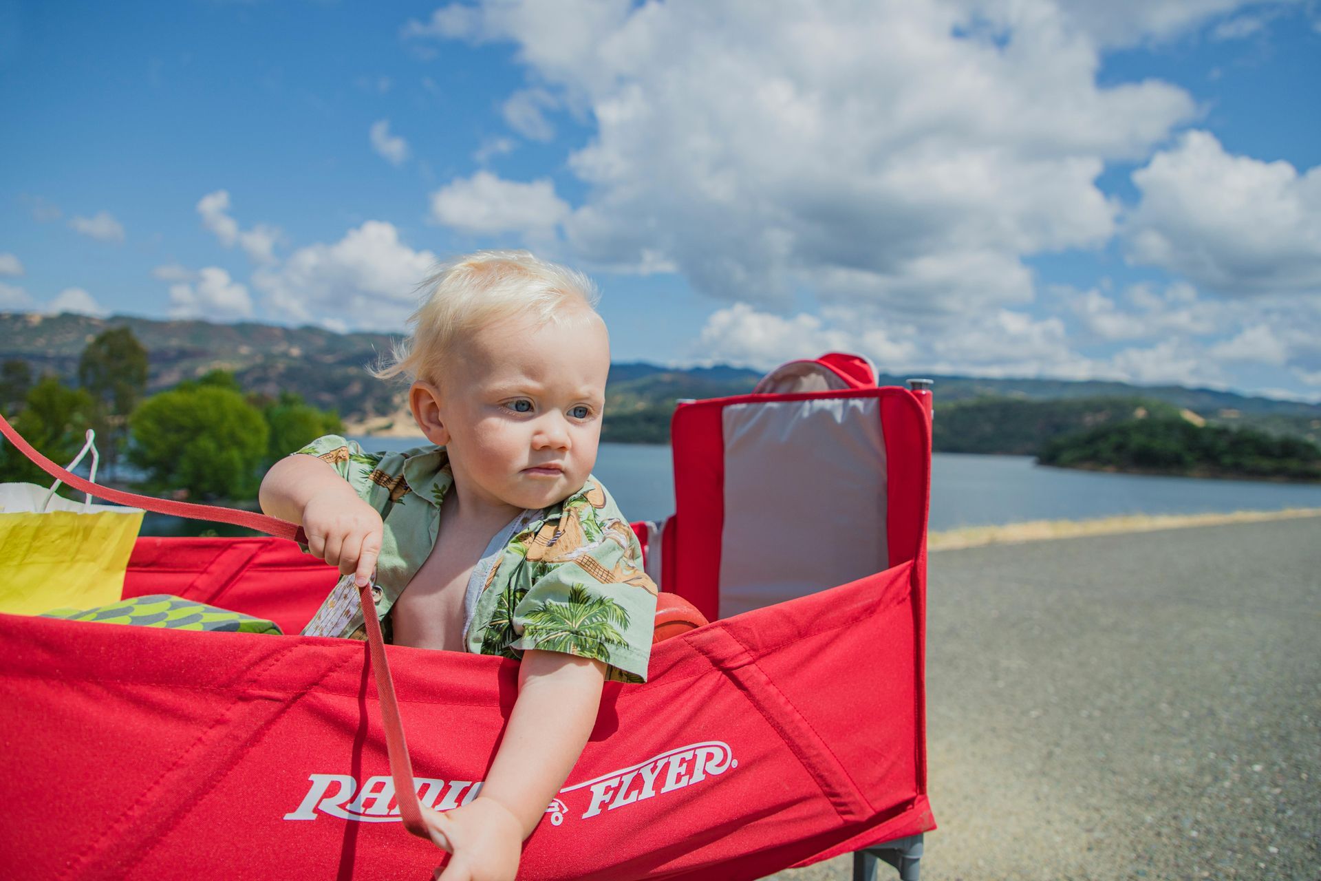 Blond toddler in a red wagon outdoors, looking to the side. Scenic lake and blue sky background.
