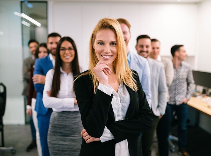 A diverse group of professionals in business attire, smiling confidently in an office setting, the blonde woman in front.