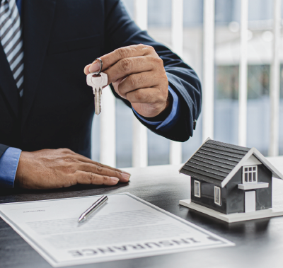 A person in a suit holds a key over an insurance document and a small house model on a desk.