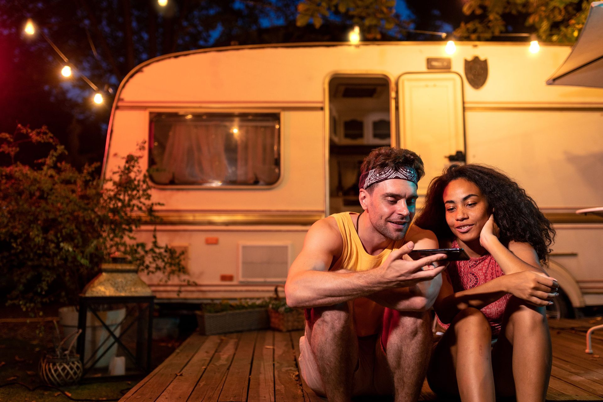 A couple sits on a wooden deck at night in front of a camper, smiling while looking at a smartphone together.