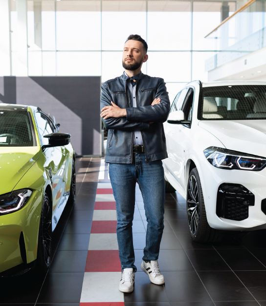 A person with a beard stands with arms crossed in a car dealership between a lime-green car and a white SUV.