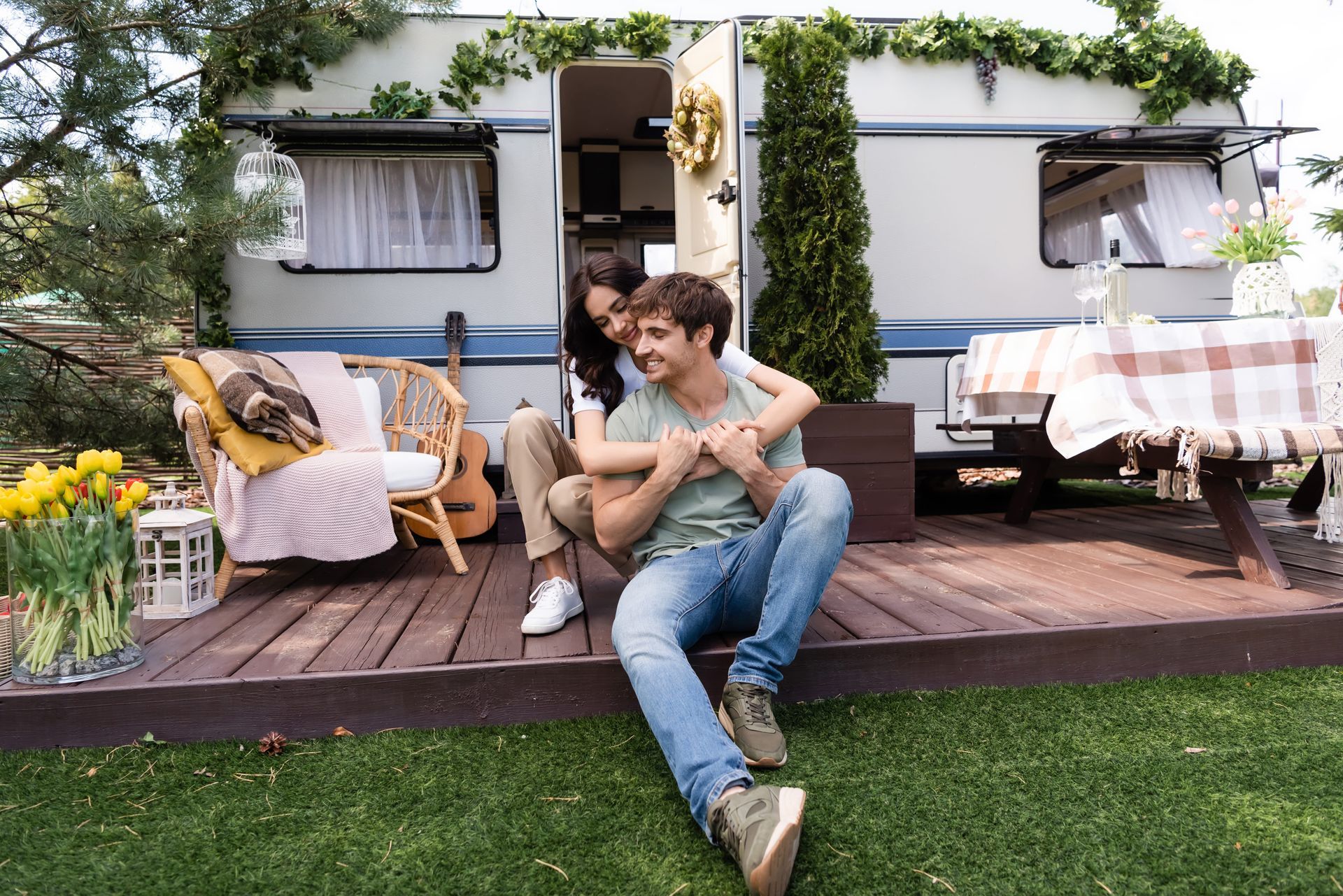 A smiling couple sits embracing on the wooden deck of a decorated camper trailer surrounded by greenery and flowers.