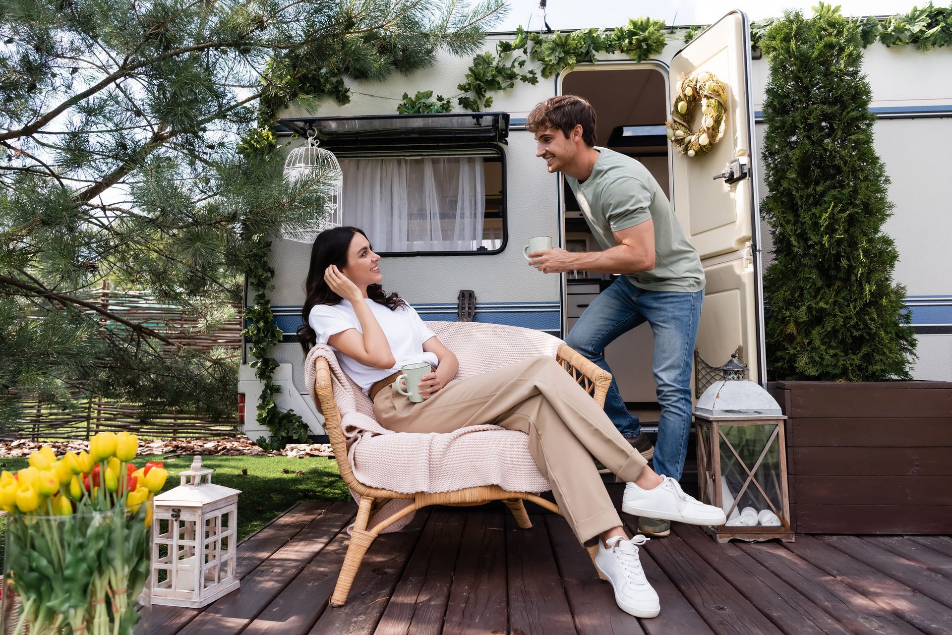 A couple chats near a vintage camper decorated with greenery, with one seated in a chair and the other standing.