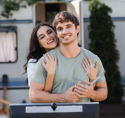 A smiling couple embraces outdoors in front of a camper, with the person in front holding grilling tongs.