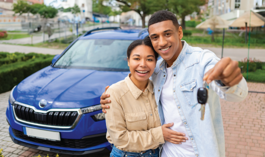 A happy couple stands in front of a blue car, smiling and holding up a set of keys to the camera.