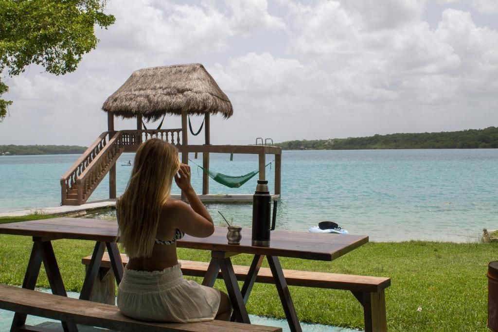 Una persona está sentada en una mesa de picnic con vistas a un lago de aguas cristalinas de color turquesa, con un muelle con techo de paja y una hamaca en el agua.