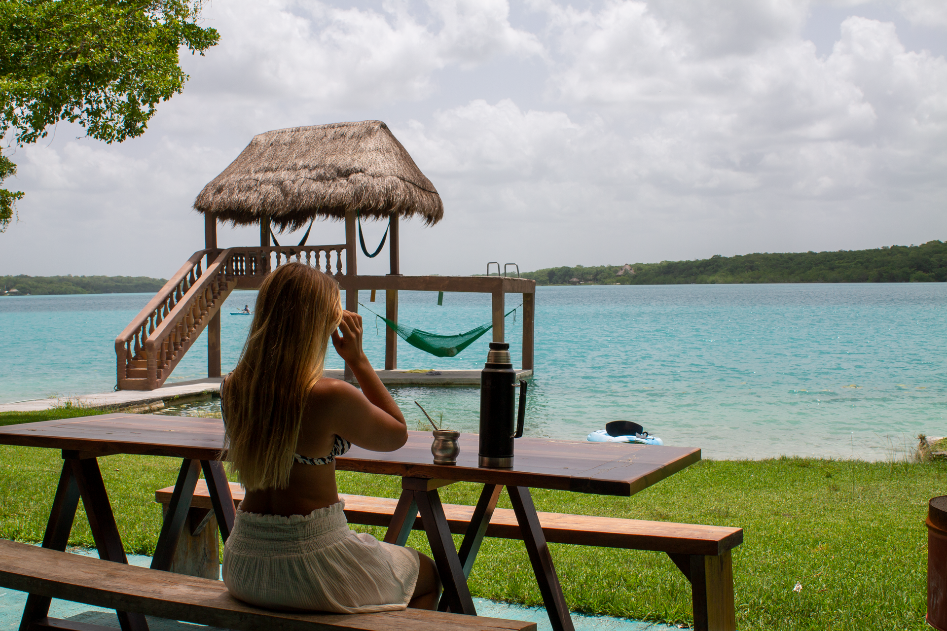 Una persona sentada en una mesa de picnic de madera con vistas a un lago de un azul intenso, con un muelle con techo de paja y una hamaca.