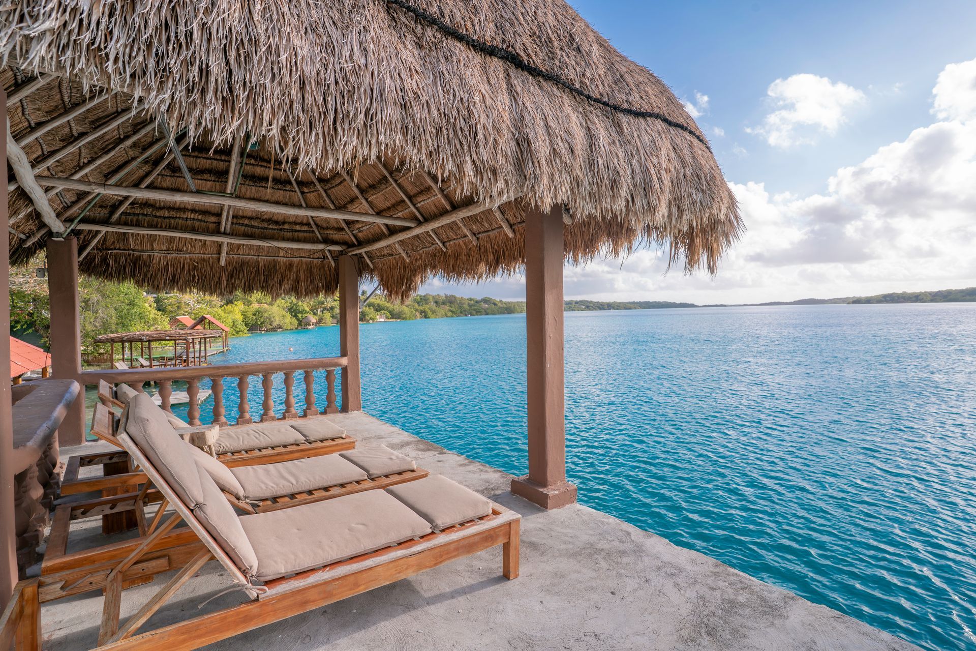 Dos tumbonas bajo una cabaña con techo de paja con vistas a una vasta extensión de agua de un azul brillante.