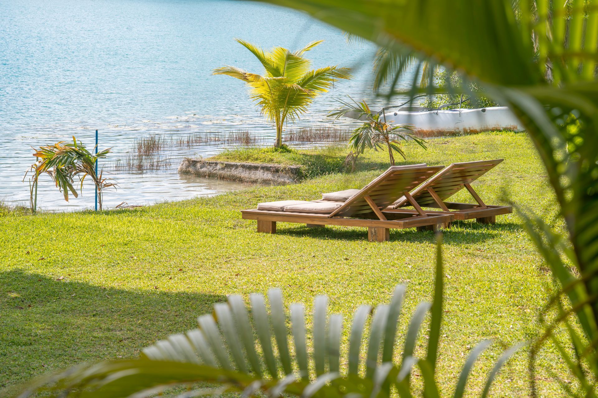 Dos tumbonas de madera sobre un césped con vistas a un lago tranquilo, parcialmente enmarcado por hojas de palmera.