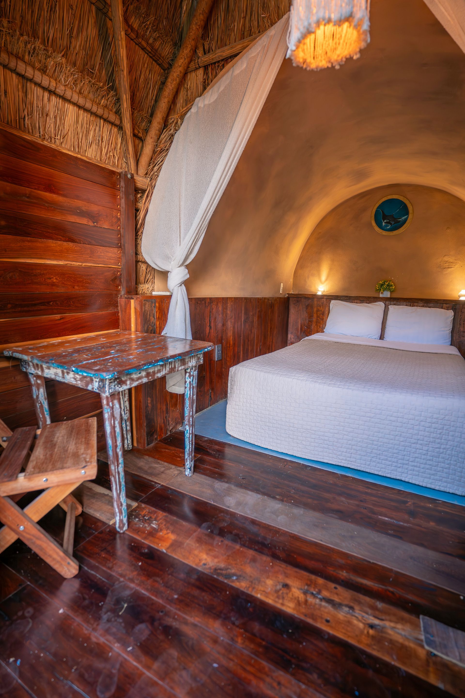 Un dormitorio acogedor con paredes de madera, una cama blanca, una mesita, una lámpara colgante y una cortina blanca.