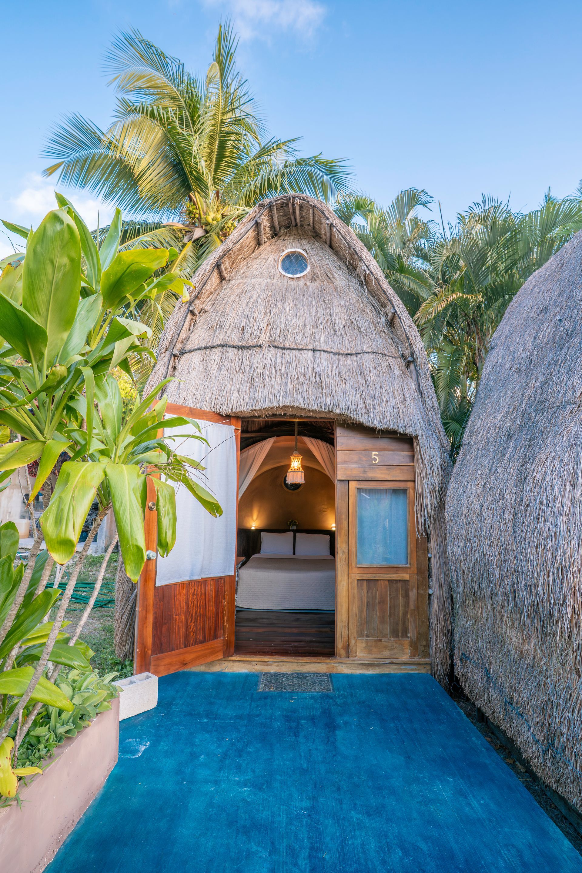 Un bungalow con techo de paja y una puerta abierta que deja ver un dormitorio acogedor e iluminado, situado en un patio azul brillante con palmeras.