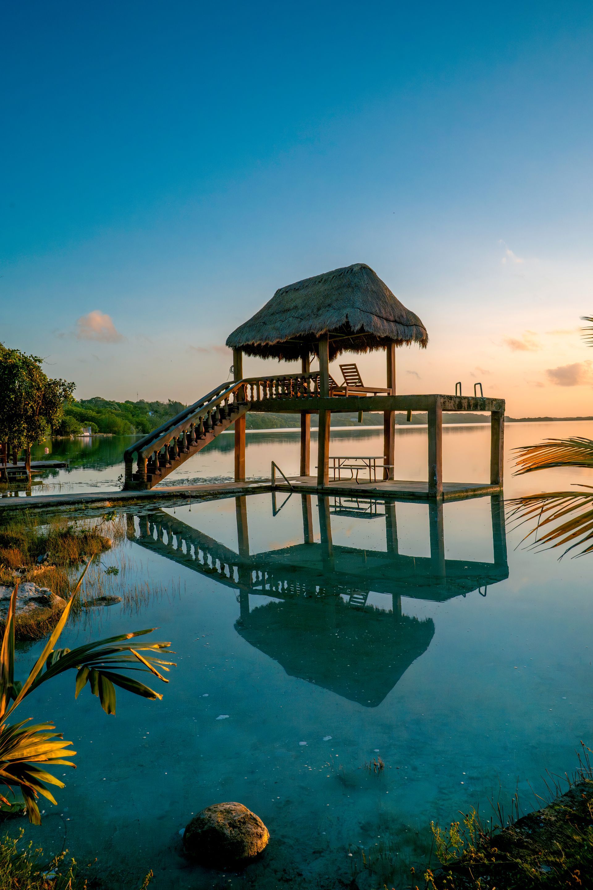 Un muelle de madera con techo de paja se alza sobre aguas tranquilas y cristalinas al atardecer, reflejándose perfectamente en la superficie.