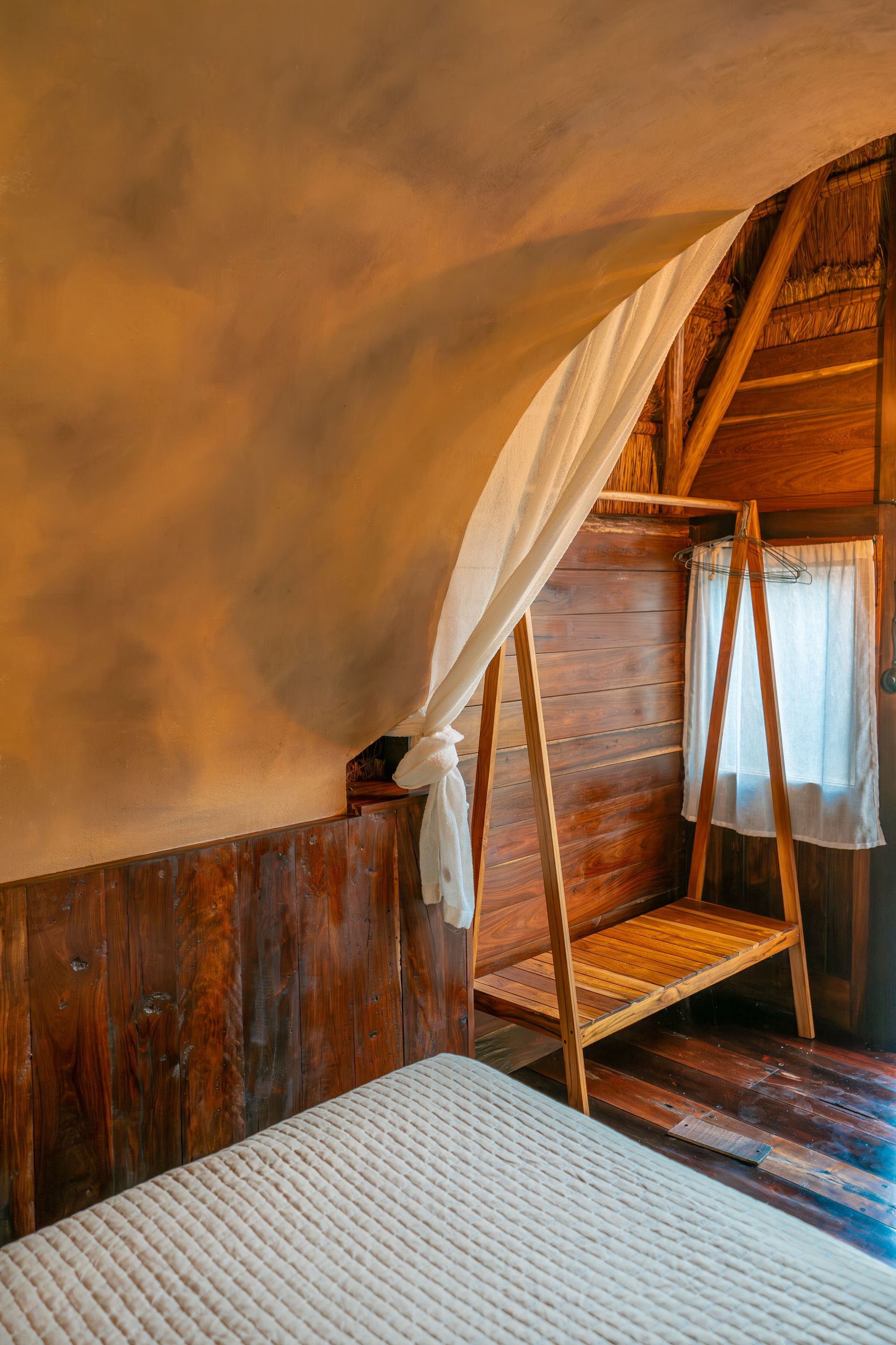 Una habitación rústica con paredes de madera, techo abovedado con textura, colchón blanco y perchero de madera con estructura abierta.