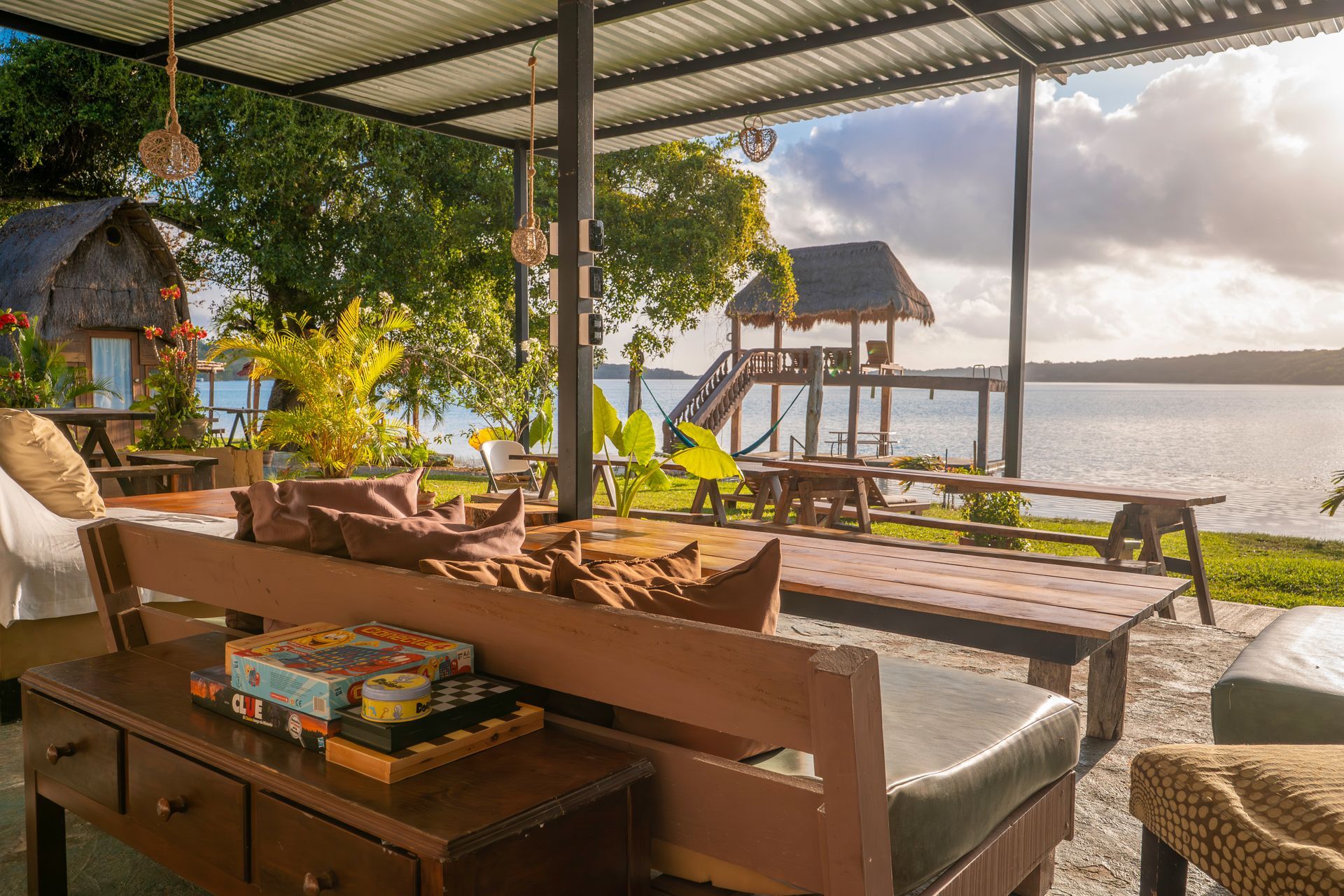 Un pabellón al aire libre con muebles de madera para relajarse, con vistas a un lago, una pequeña cabaña y un muelle bajo un cielo despejado.