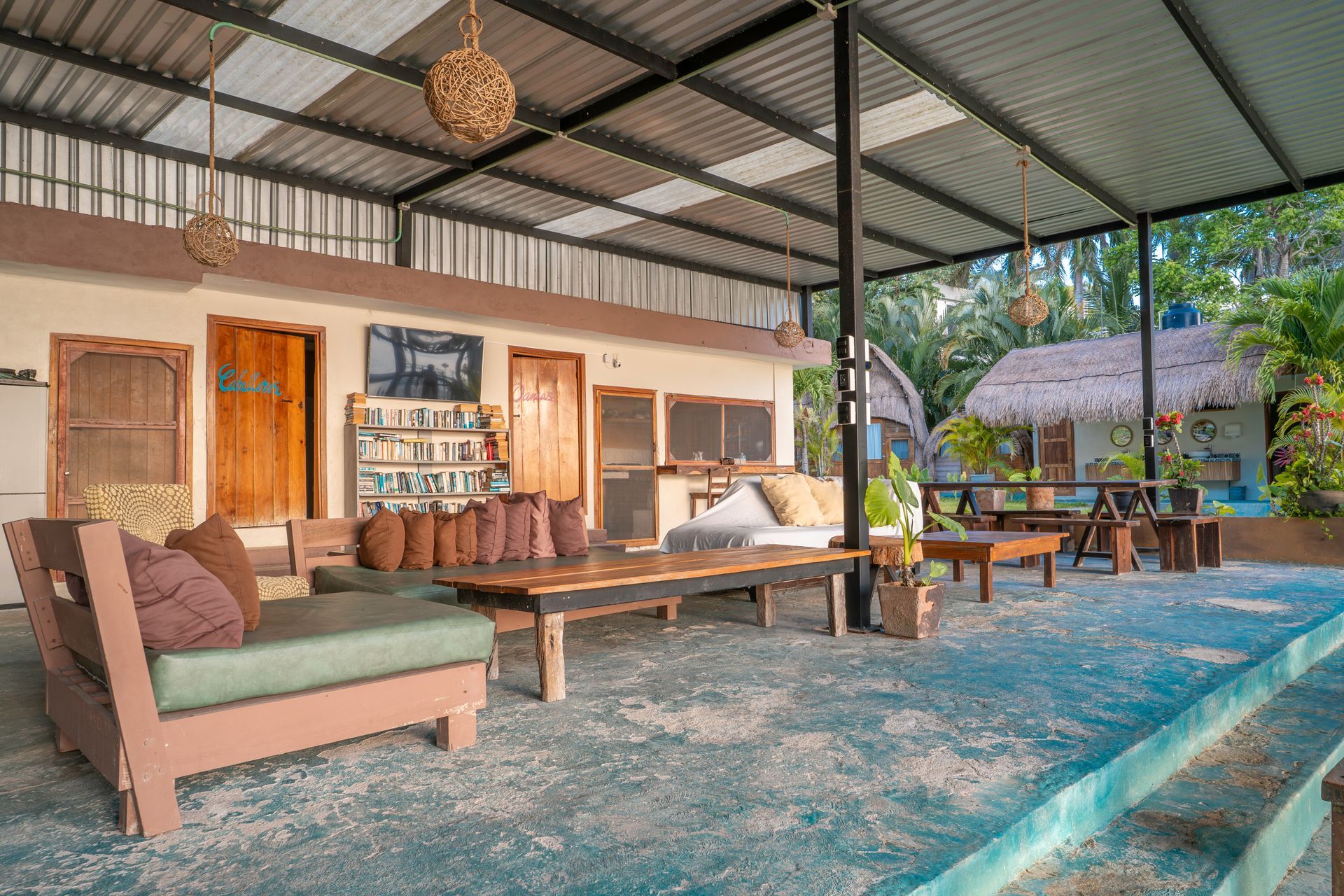 Un patio exterior cubierto cuenta con bancos de madera con cojines y una mesa central, todo ello sobre un suelo de hormigón pintado de azul.