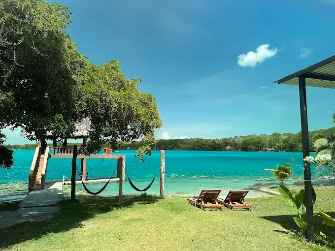 Un complejo turístico a orillas del lago, rodeado de césped, con dos tumbonas, un muelle de madera y hamacas bajo un árbol, con vistas a aguas turquesas.