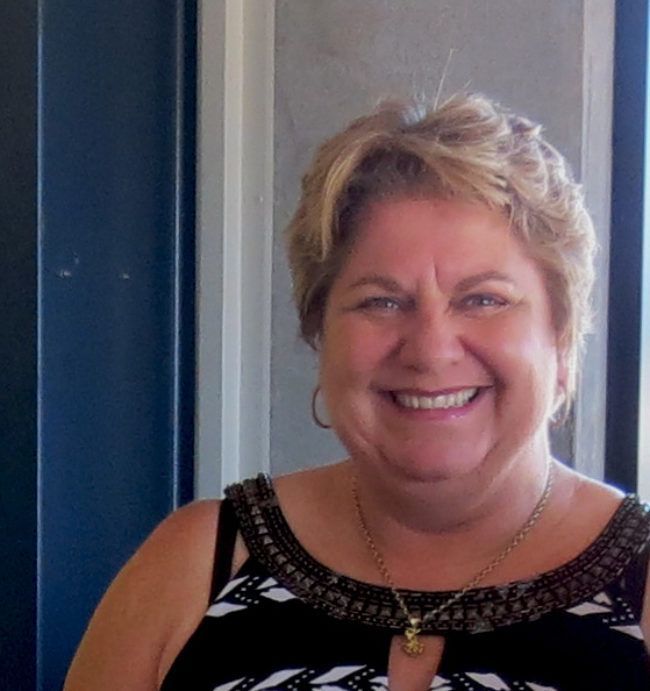 Smiling woman with blonde hair, wearing a black and white patterned top, gold necklace, standing outdoors.
