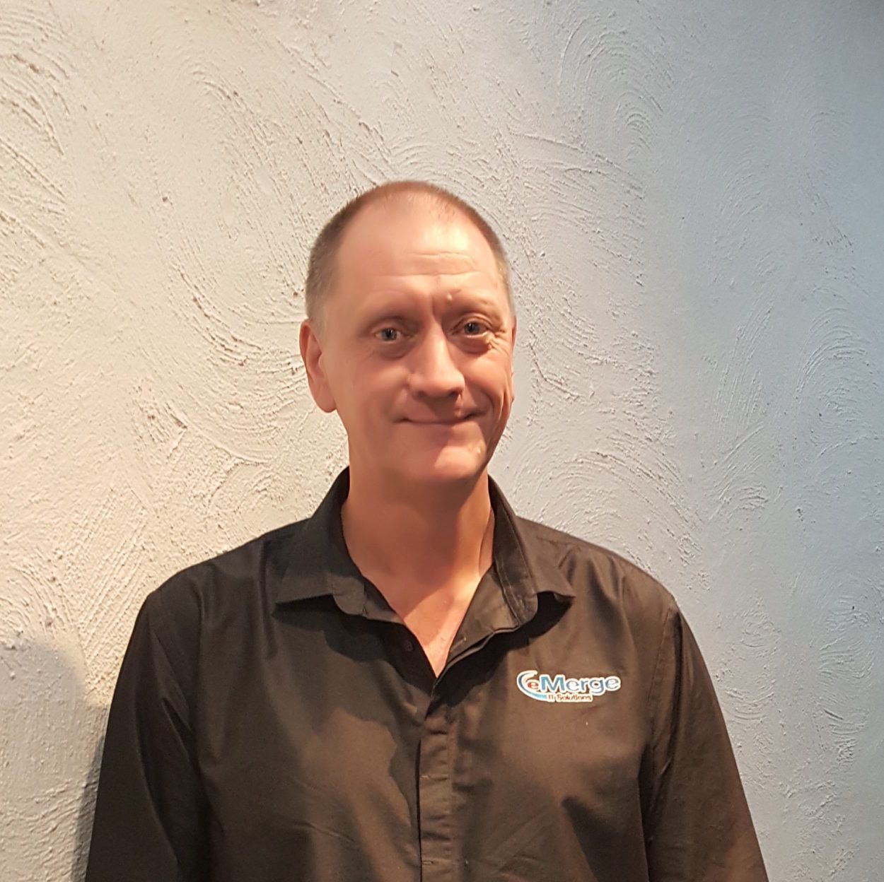 Man with fair skin and thinning hair, smiling in a black shirt, standing in front of a textured wall.