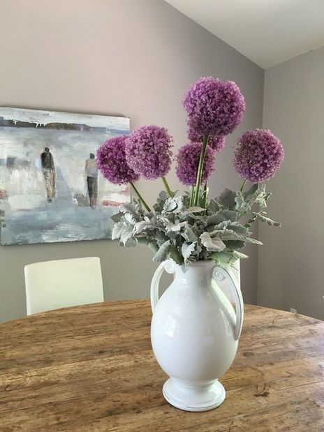 A white vase filled with purple flowers is sitting on a wooden table.