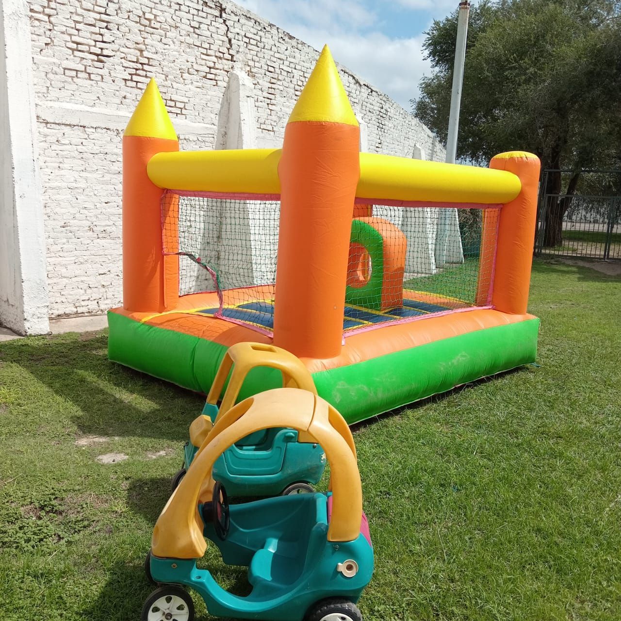Castillo hinchable colorido sobre el césped junto a una pared de ladrillos blancos, con un pequeño coche de juguete delante.