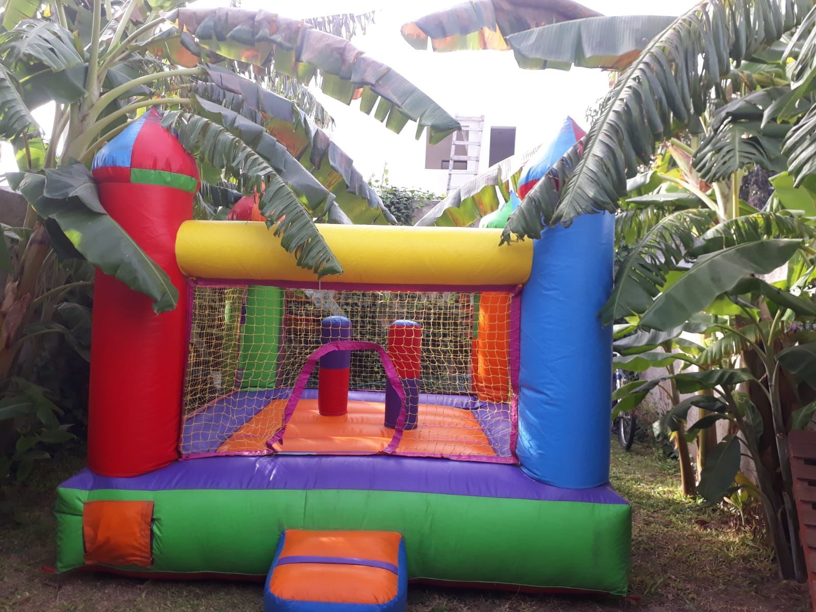 Castillo hinchable colorido al aire libre con dos niños dentro, rodeado de plantas tropicales.