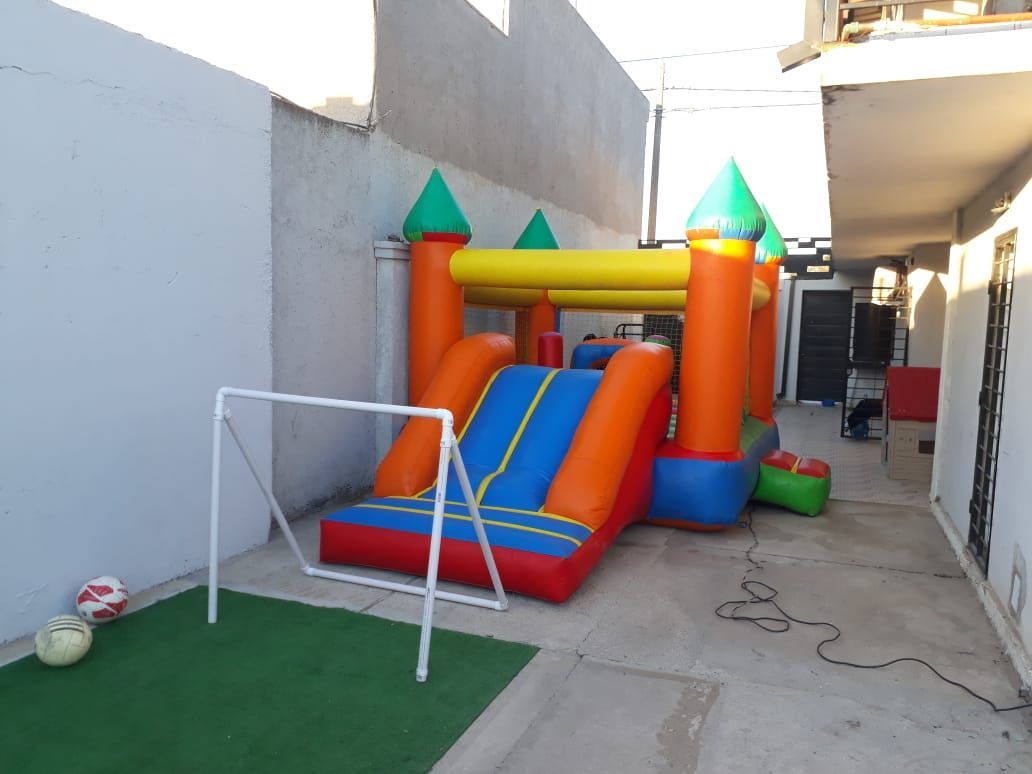 Castillo hinchable colorido en un estrecho patio exterior con una pequeña portería de fútbol y césped artificial.
