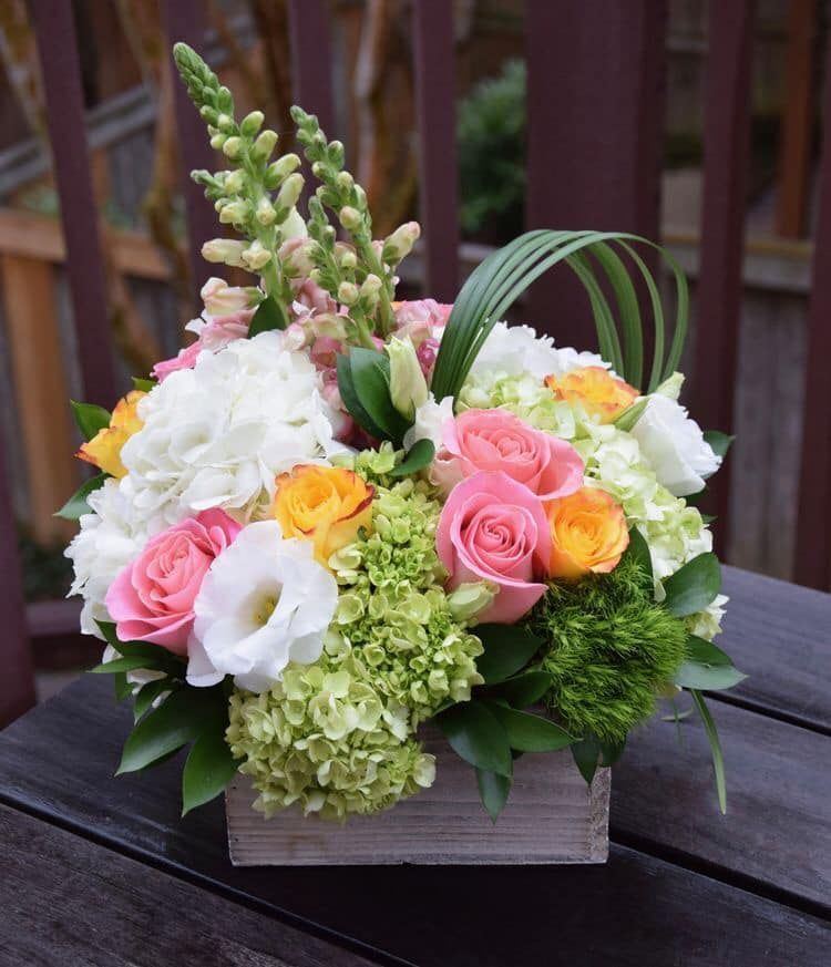 A floral arrangement in a wooden box featuring pink roses, white hydrangeas, orange roses, and snapdragons.