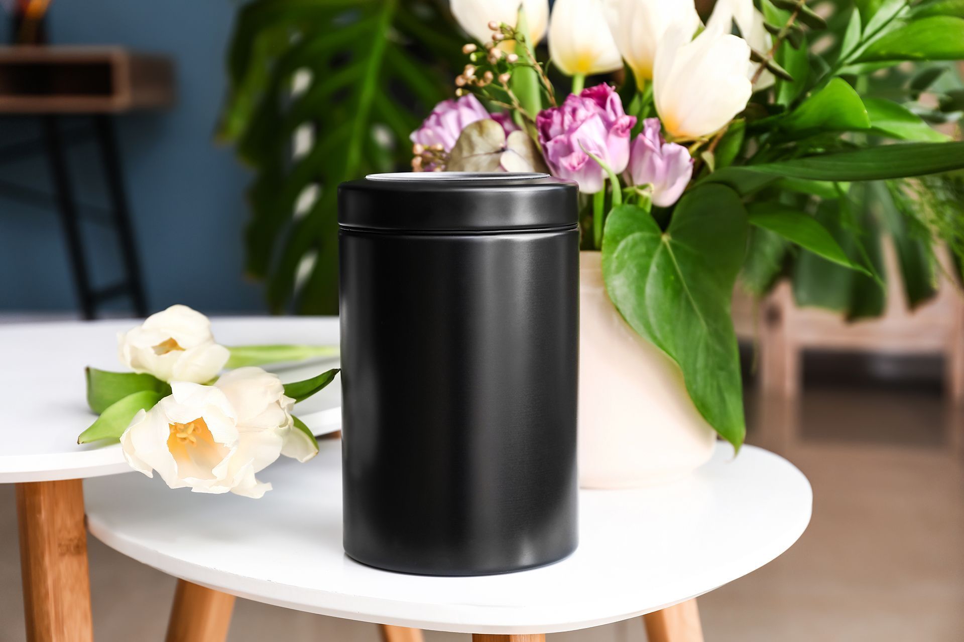 A black cylindrical cremation urn sits on a white table next to a vase of tulips.