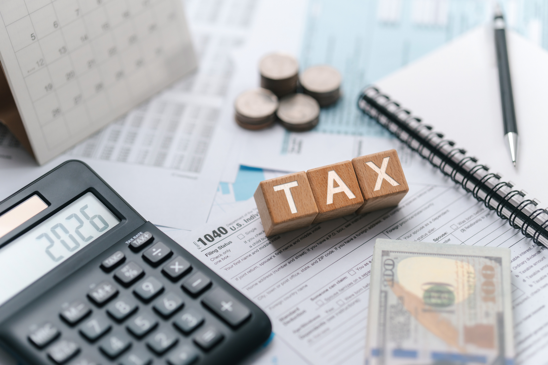 Calculator, tax forms, cash, and wooden “TAX” blocks on a desk with a notebook and pen.