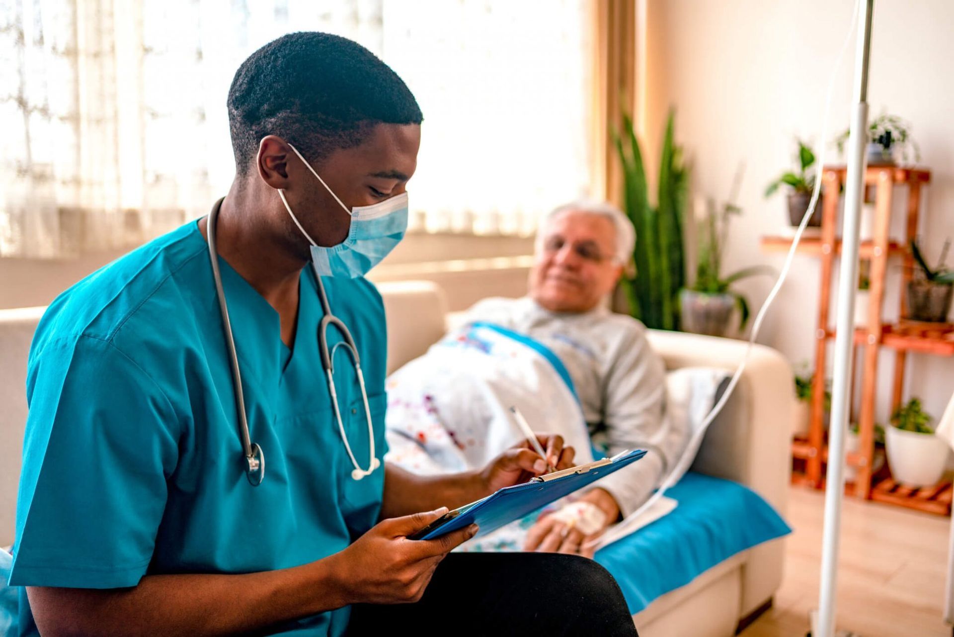 Nurse in blue scrubs with a stethoscope, wearing a mask, taking notes. Elderly patient rests in bed.