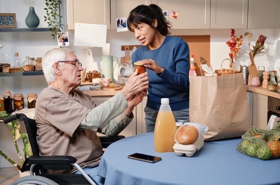 Woman assists older person in a wheelchair, opening a jar in a kitchen, groceries on table.