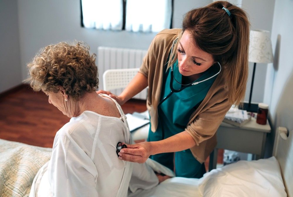 Nurse using a stethoscope to listen to the back of a patient in a bedroom.