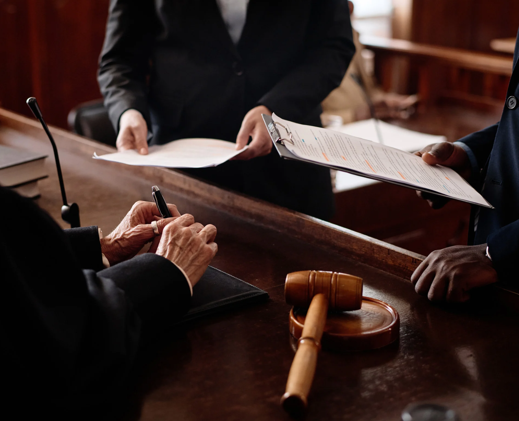 Judge in courtroom, signing papers; lawyers stand nearby, holding documents.