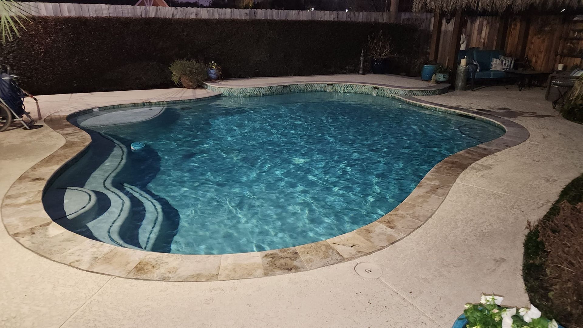 Nighttime view of a turquoise pool with stone coping and a paved surround in a backyard.