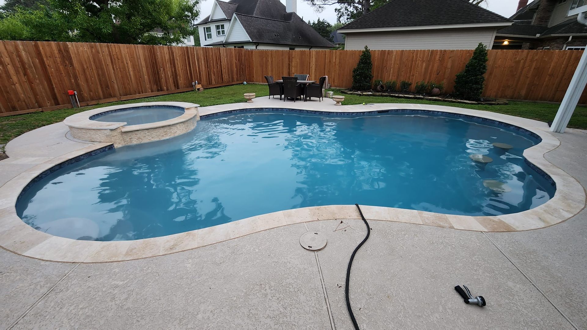 Backyard pool with spa, dining set, and wood fence. Concrete patio, blue water, and green grass.