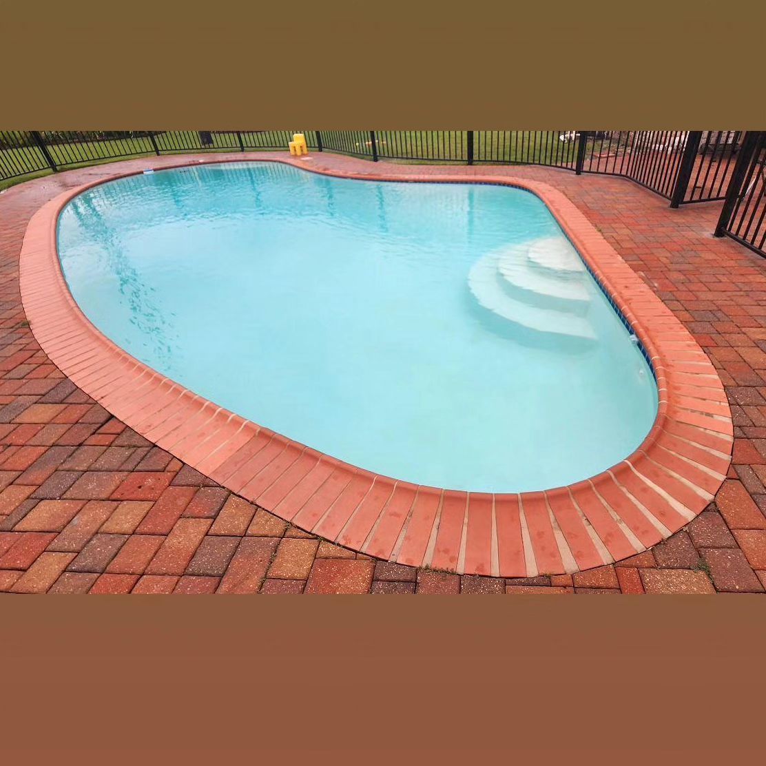 Oval swimming pool surrounded by red brick, with white steps. The water is light blue, set outdoors.