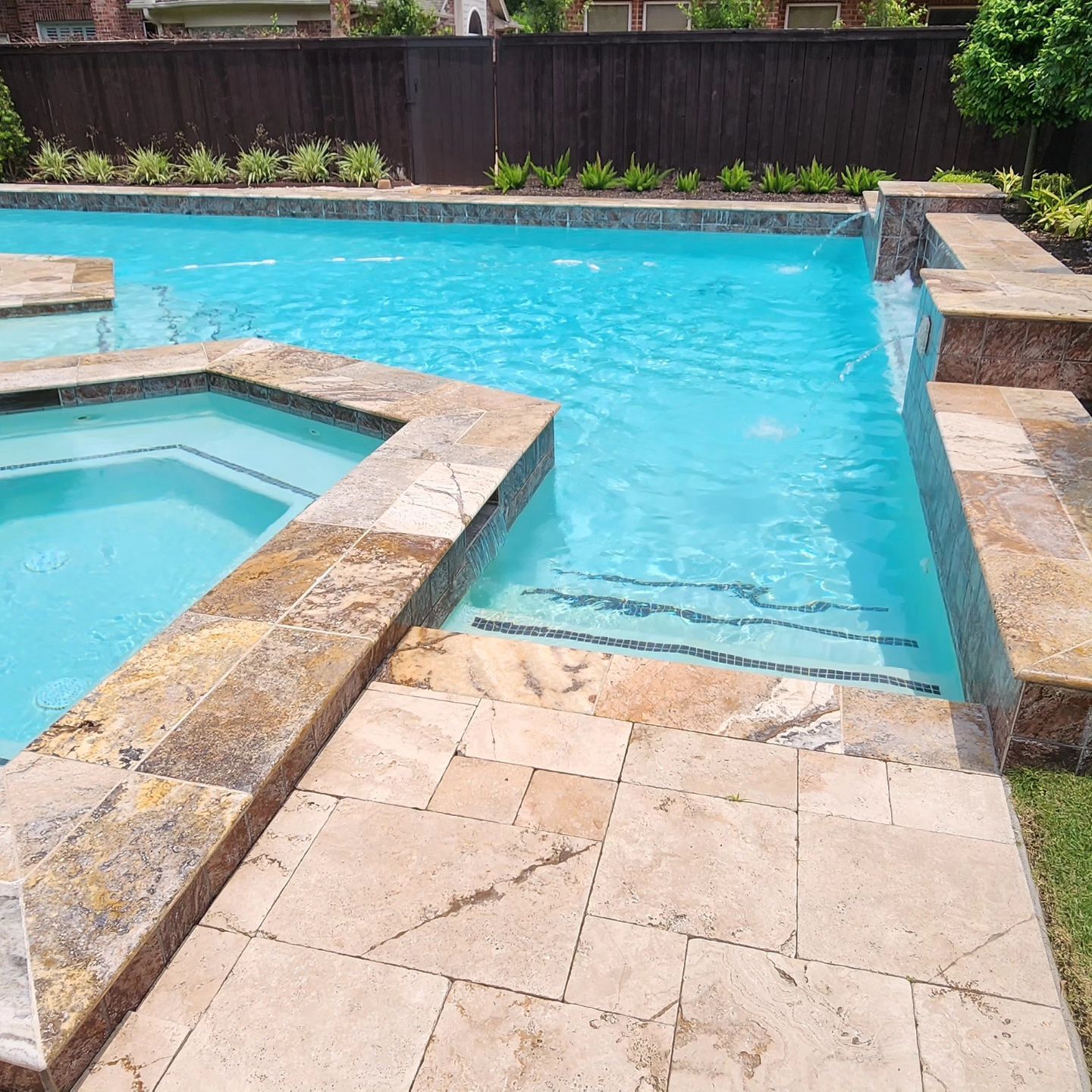 A swimming pool with a connected jacuzzi and stone patio. Clear blue water.