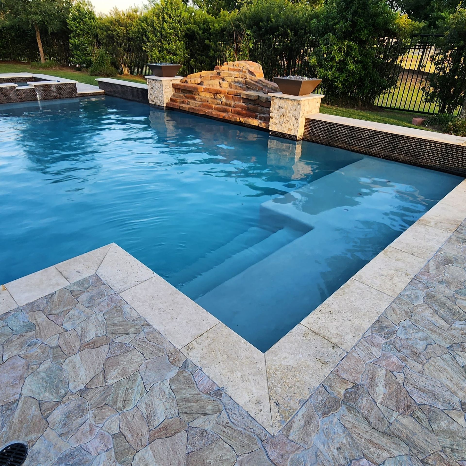 Swimming pool with stone deck, steps, and rock waterfall. Blue water, sunny outdoor setting.