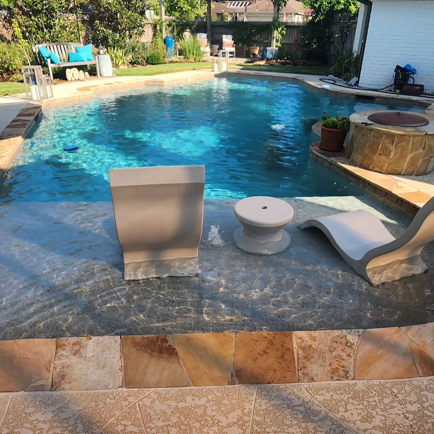 Pool with two chairs and a side table on the deck. Blue water, stone surround.