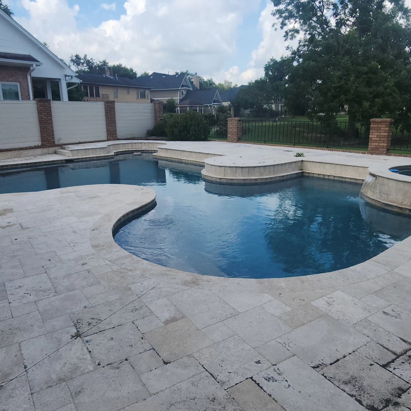 A swimming pool with light-colored stone surround. Sky and trees are in the background.