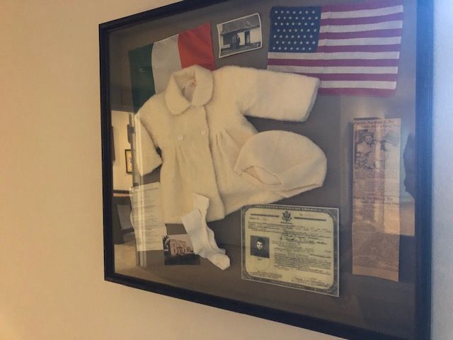 Framed display with Irish and US flags, baby clothes, and documents. Light-colored outfit, passport, and other memorabilia.